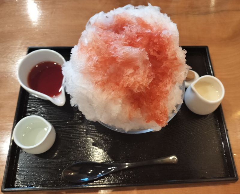 A fluffy bowl of shaved ice topped with strawberry syrup, served with condensed milk and clear syrup on a black tray at Asami Reizo, a famous shaved ice café in Nagatoro, Japan.